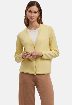 Geel vest met een V-hals, vijf knopen en lange mouwen. De stof lijkt zacht en lichtgewicht, gecombineerd met beige broeken.