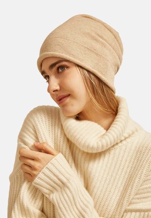 Beige gestrickte Mütze mit weicher Textur, für einen engen Sitz entworfen. Getragen mit einem cremefarbenen, gerippten Rollkragensweater.