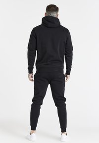 Black sweatshirt with a hood and elastic cuffs, paired with matching black jogger pants. Smooth texture, fitted design, white sneakers.