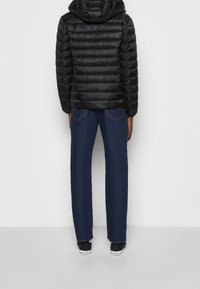 Sort polstret jakke med hætte, parret med mørkeblå lige ben denim jeans og sorte sneakers, set fra bagsiden.