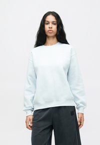 Γυναίκα με μακριά μαύρα μαλλιά, φορώντας ένα ανοιχτό μπλε φούτερ και σκούρες γκρι παντελόνες, σε ένα απλό λευκό φόντο.