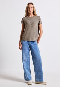 T-shirt loose en vert olive avec des manches courtes, ornée de détails de couture diagonale, associée à un jean clair à jambes larges.