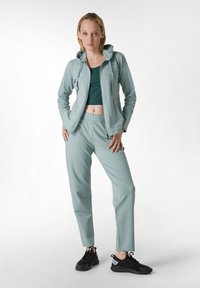 Giacca sportiva verde chiaro con cappuccio regolabile, abbinata a una maglia coordinata e pantaloni a sigaretta. Scarpe nere completano l'outfit.