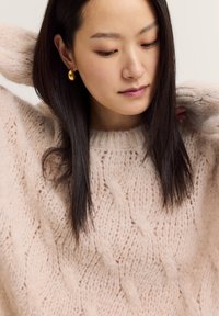 Helles rosa Kabelstrickpullover mit Rundhalsausschnitt und strukturierten Mustern. Das Model hat lange, dunkle Haare und trägt goldene Creolen.