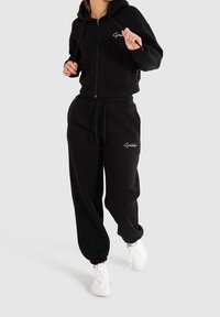 Felpa nera con zip, maniche lunghe e tasche frontali, abbinata a pantaloni jogger neri con vita elastica. Entrambi realizzati in un morbido tessuto testurizzato.