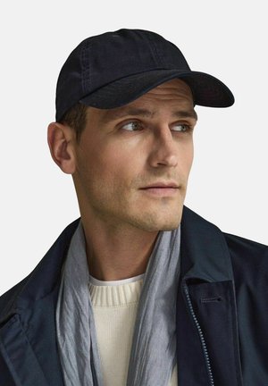 Mørkeblå baseballcap med buet skygge, lavet af stof. Båret med et gråt tørklæde og en cremefarvet sweater, sat mod en hvid baggrund.