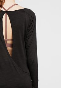 Haut à manches longues noir avec un design à dos ouvert et des fines bretelles superposées de couleur mauve, avec une texture douce et lisse.