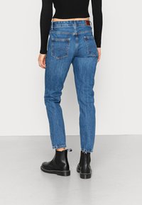 Jeans de ganga azul com um ajuste justo, cintura média e dois bolsos traseiros. Usados com botas chelsea pretas e uma camisola preta canelada.