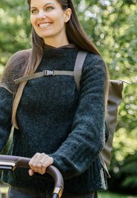 Nainen hymyilee työntäessään lasta kärryä ulkona, päällään tumma neule ja reppu, ympärillään vihreää kasvillisuutta.