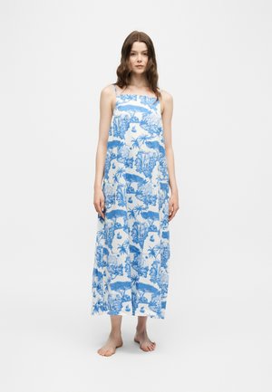 Vrouw staat blootsvoets en draagt een witte mouwloze maxidress met blauwe tropische en dierenprints.