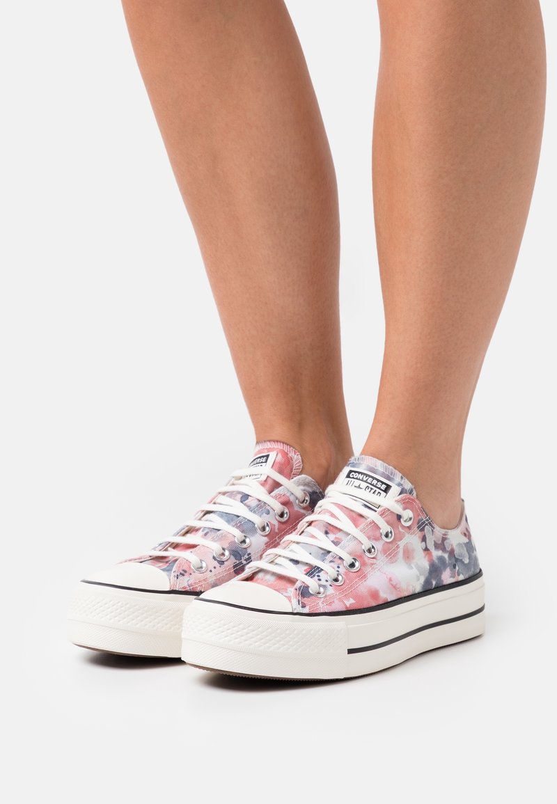 Pieds chaussés de baskets Converse basses avec un motif tie-dye rose, gris et blanc et des semelles en caoutchouc blanc épais avec des rayures noires.