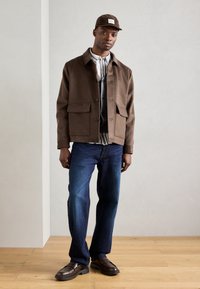 Giacca in misto lana marrone con due tasche frontali, indossata sopra un cardigan grigio e una camicia a righe, abbinata a jeans in denim blu e scarpe marroni.