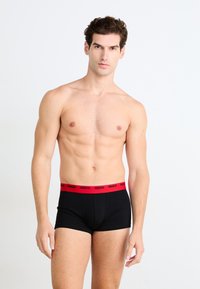 Schwarze Boxershorts mit rosem elastischen Bund, auf dem "HUGO" in fetten Buchstaben steht. Das Modell steht aufrecht vor einem neutralen Hintergrund.