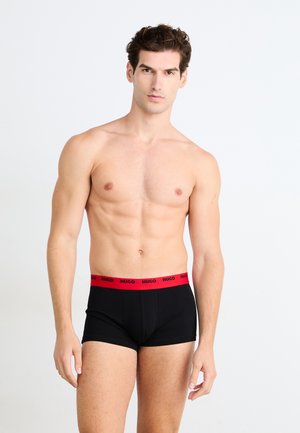 Zwarte boxershorts met een rode elastische tailleband waarop "HUGO" in vette letters staat. Het model staat rechtop, met een neutrale achtergrond.