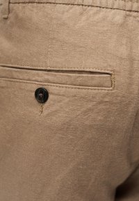 Τσέπη παντελονιού από μπεζ ύφασμα με μία μαύρη πλακέ κουμπί και ορατές ραφές.