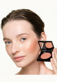 Blush compact with a pinkish peach shade, housed in a sleek black case. Model holds the compact near her cheek, showcasing product application.