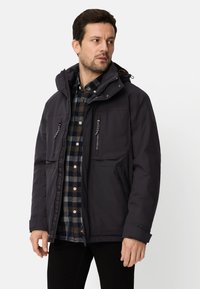 Czarna ocieplana kurtka z zamkiem błyskawicznym z przodu, regulowanym kapturem i wieloma kieszeniami. Noszona na koszuli w kratę, o gładkiej fakturze.
