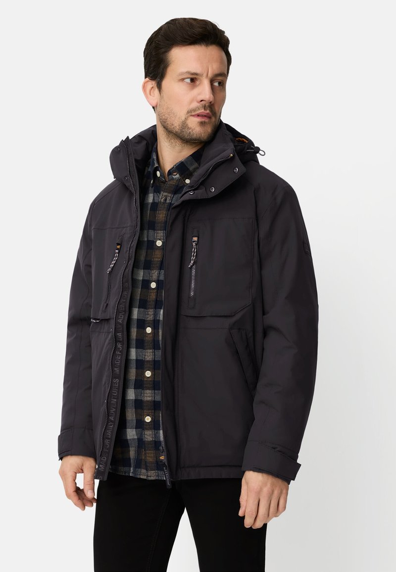 Czarna ocieplana kurtka z zamkiem błyskawicznym z przodu, regulowanym kapturem i wieloma kieszeniami. Noszona na koszuli w kratę, o gładkiej fakturze.