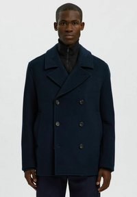 Pardessus in lana blu navy con ampi rever, doppio petto e due tasche laterali. Indossato su un capo nero con collo alto e zip.