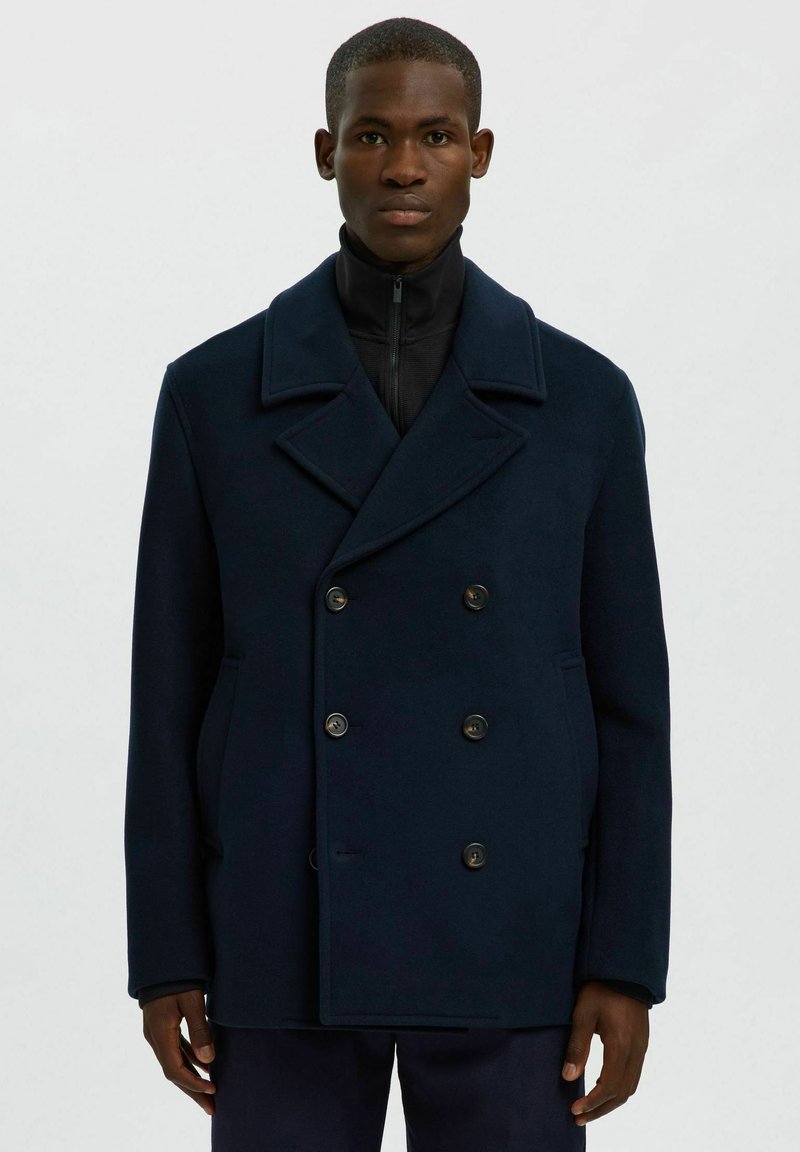Pardessus in lana blu navy con ampi rever, doppio petto e due tasche laterali. Indossato su un capo nero con collo alto e zip.