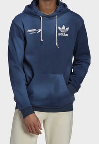 Námořnická modrá mikina s kapucí a přední kapsou typu klokanka, se stahovacími šňůrkami a bílým logem a brandingem Adidas. Měkká textura a ležérní design.