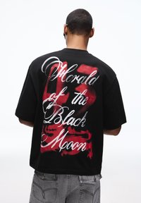 Fekete, túlméretezett póló, amelynek hátoldalán piros és fehér, graffiti stílusú felirat található, laza szabású és rövid ujjú.