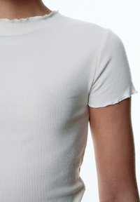 Witte geribbelde t-shirt met korte mouwen, voorzien van een gestructureerde afwerking en golvende halslijn. De stof lijkt rekbaar en sluit goed aan op het lichaam.