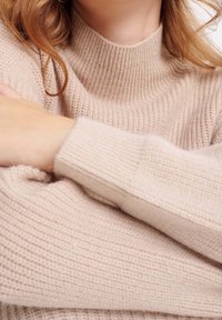 Pull beige en maille avec un col roulé côtelé, un tissu texturé et des manches longues avec des poignets côtelés, offrant un aspect chaleureux.