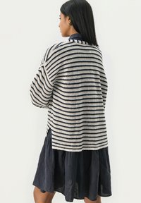 Stripet sweater i creme og marineblå, strikket tekstur, oversized pasform, åbne sider, parret med en marineblå linnedkjole. Casual lag-på-lag design.