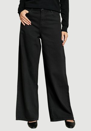 Femme portant un pantalon noir taille haute à jambes larges avec poches avant et des chaussures noires à bout pointu sur un fond uni.