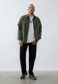 Chemise à boutons avec un motif écossais vert sur un t-shirt blanc, associé à un pantalon noir et des chaussures de sport ; style décontracté avec un design superposé.