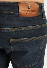 Tmavě modré džíny z denimu s texturovaným povrchem, s hnědou koženou nášivkou na pásku, oranžovým šitím a zadními kapsami s zakřivenými švy.