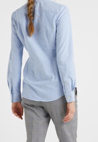 Vaaleansininen pitkähihaista paitaa, joka on valmistettu sileästä kankaasta, pieni kaulus, napilliset hihansuut ja muotoiltu malli sivusaumoilla.