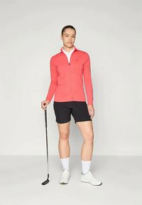 Koralfarvet zip-up langærmet trøje, hvid krave, sorte shorts, holder en golfkølle, iført hvide sko og sokker, stående på en lys baggrund.