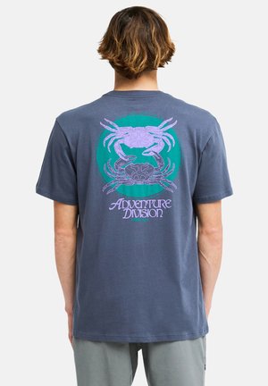 Osoba o krótkich brązowych włosach nosi ciemnoniebieski t-shirt z dwoma fioletowymi krabami i napisem "Adventure Division" na plecach.