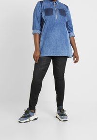 Tunika in denim blu chiaro con colletto zip e due tasche sul petto, abbinata a jeans skinny neri e sneakers multicolori.