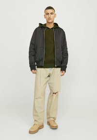 Zwarte bomberjack, groene hoodie en lichtbeige gescheurde jeans. Over het algemeen een relaxte pasvorm met ribgebreide cuffs en tan schoenen. Minimalistisch ontwerp.