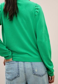 Vihreä neuletakki, jossa on ritilävarsi ja helma, sivuilla rypytystä ja tähkäkuvio vasemmassa hihassa. Yhdistetty vaaleansinisille farkkushortsille.