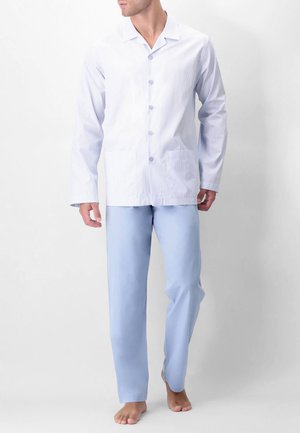 Uomo in piedi a piedi nudi che indossa pantaloni azzurri e una camicia bianca a maniche lunghe con sottili righe e tasche anteriori.