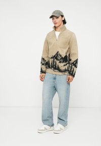 Beige fleece-tröja med halvdragkedja, med en svart landskapsdesign av fjäll och träd vid fållen; matchad med ljusblå jeans och vita sneakers.