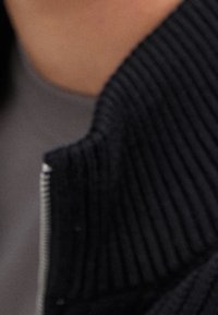 Black zip-up collar with ribbed texture, layered over a gray garment. Close-up detail focuses on fabric and stitching.