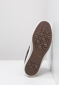 Semelle en caoutchouc brun d'une basket avec un motif texturé, disposant d'une semelle intermédiaire blanche et d'une tige en toile colorée avec des œillets en métal.