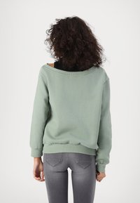 Sudadera verde claro de corte holgado, con diseño off-the-shoulder, puños y cinturilla de canalé. Lleva puesta sobre una camiseta negra, combinada con jeans grises.
