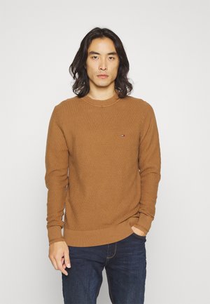 Maglione marrone lavorato a maglia con texture a coste e scollatura a girocollo. Presenta un piccolo logo sul petto, abbinato a jeans blu scuro.
