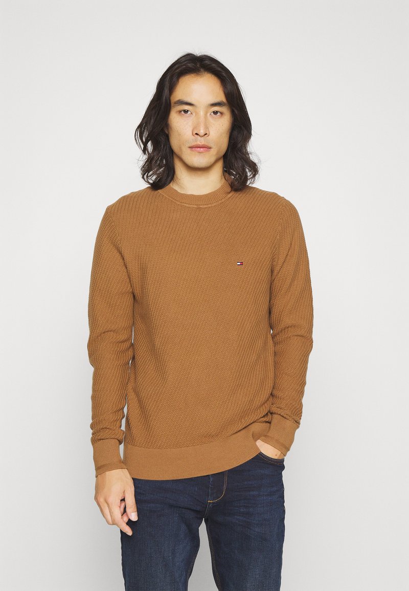 Maglione marrone lavorato a maglia con texture a coste e scollatura a girocollo. Presenta un piccolo logo sul petto, abbinato a jeans blu scuro.