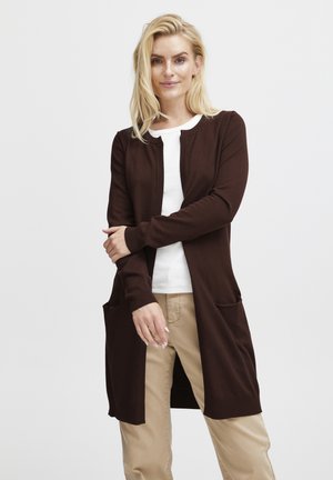 Brauner, langer Cardigan aus weichem Strickstoff, mit Rundhalsausschnitt, gerippten Bündchen und zwei vorderen Taschen. Über einem weißen Hemd getragen.