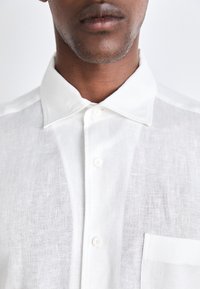 Camisa de lino blanca con cuello clásico, que presenta un bolsillo frontal y un botón en la parte delantera. Textura suave con un diseño ligero y transpirable.