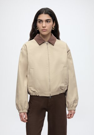Frau mit langen dunklen Haaren, die eine beige Jacke mit Reißverschluss und braunem Kragen und braune Hose trägt, steht vor einem weißen, schlichten Hintergrund.