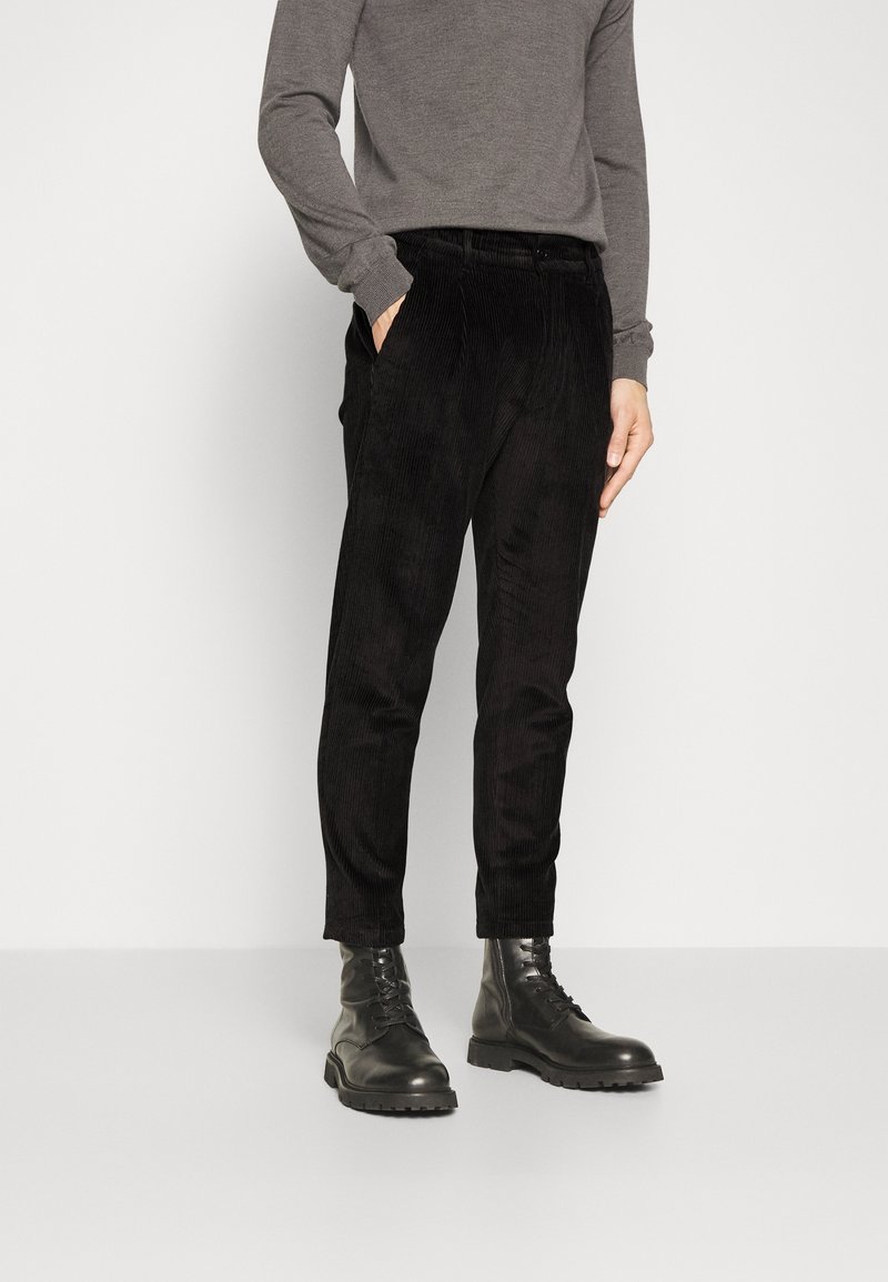 Pantalon en velours côtelé noir avec une coupe droite, doté de poches latérales et d'une surface texturée, associé à des bottes noires à lacets.