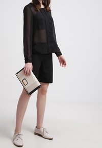 Donna in blusa nera a maniche lunghe e pantaloni corti neri, che tiene una borsa beige, indossando scarpe beige con lacci su sfondo bianco.